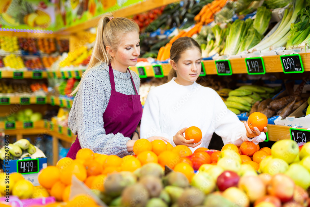 Friendly saleswoman helping teenage girl to choose ripe sweet oranges in fruit and vegetable section of supermarket..