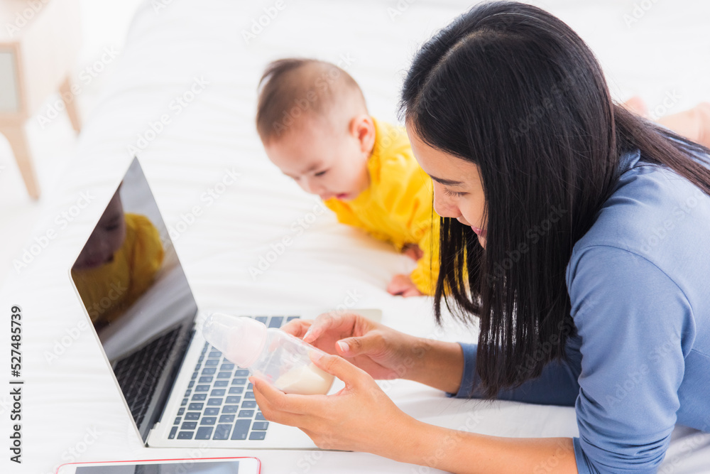 Asian new young beautiful mother finding and reads information on a laptop for milk mixing on a bottle in the bedroom at home Asian new young beautiful mother finding and reads information on a laptop for milk mixing on a bottle in the bedroom at home