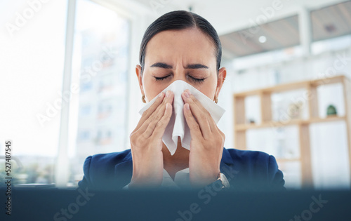 Covid, virus and sick with a business woman blowing her nose with a tissue and sneezing while working in her office. Allergies, sneeze and flu with a female employee suffering from a cold at work