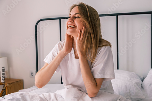 Waking up in the morning. Young happy woman smiling and touching her face sitting in bed with her eyes closed after waking up. Morning bliss. Healthy sleep, body recovery