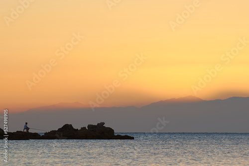 A man is fishing by the mediterranean sea with the rising morning sun in the background © Stefan