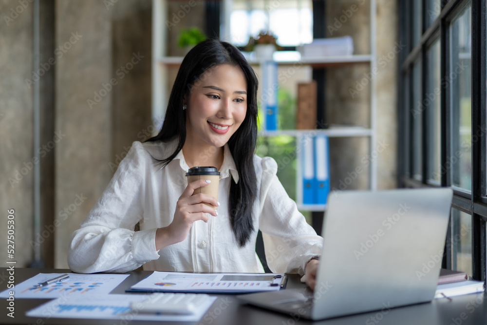 Pretty Asian businesswoman sitting happily working on her laptop and drinking coffee smiling and laughing brightly in the office. Pretty Asian businesswoman sitting happily working on her laptop and drinking coffee smiling and laughing brightly in the office.