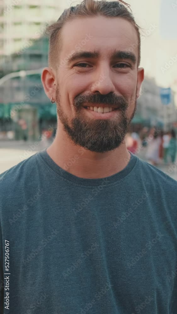 VERTICAL VIDEO: Young smiling man with beard standing in front of busy intersection VERTICAL VIDEO: Young smiling man with beard standing in front of busy intersection