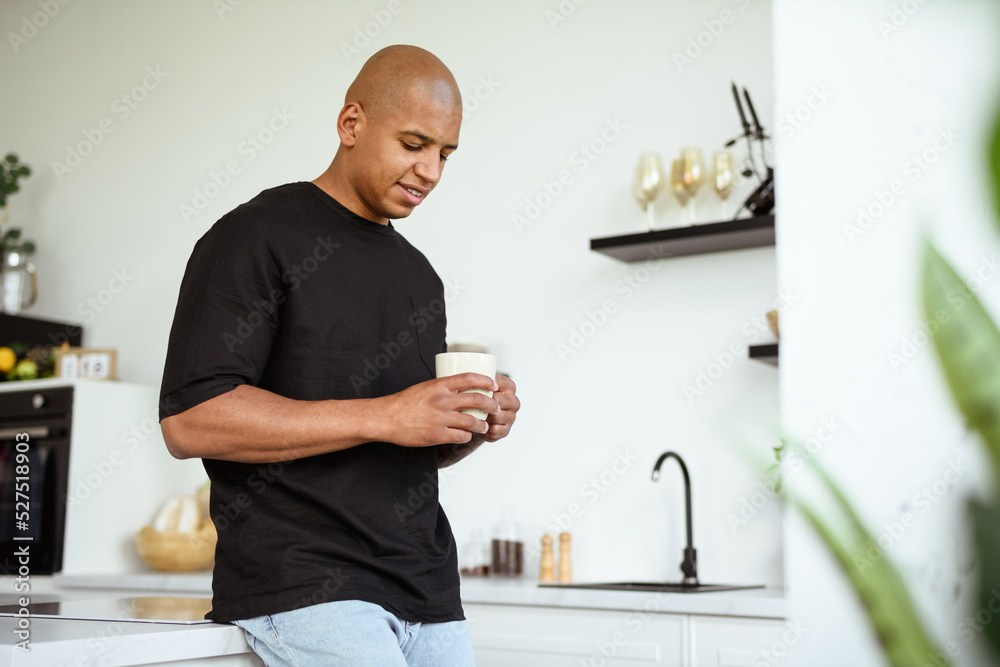Positive african american man in black t-shirt holding cup of coffee in kitchen Positive african american man in black t-shirt holding cup of coffee in kitchen