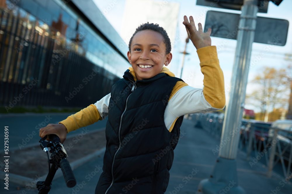 Φωτογραφία Happy black teen boy waving hand doing hi gesture greeting his friend, driving scooter on city streets, going to skate park to train new skills and tricks