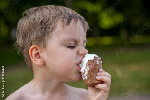 A cute blond boy appetizingly eats ice cream in the summer  sitting on the bank of the river. Cool off by the water. Funny facial expression.