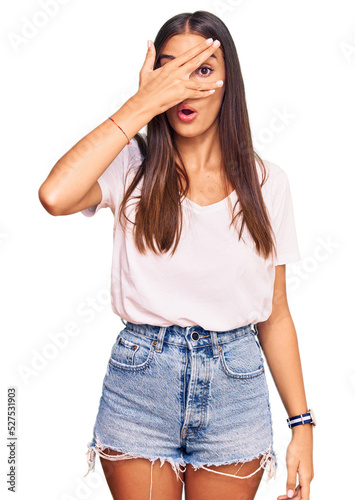Young hispanic woman wearing casual white tshirt peeking in shock covering face and eyes with hand, looking through fingers with embarrassed expression.