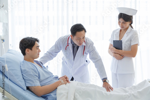 Senior doctor and young male patient who lie on the bed while checking symptom, consult and explain with nurse taking note and supporting in hospital wards.
