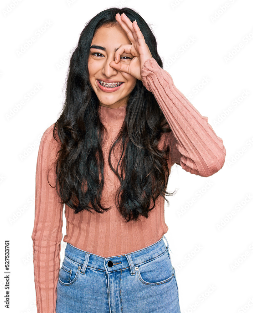 Hispanic teenager girl with dental braces wearing casual clothes doing ok gesture with hand smiling, eye looking through fingers with happy face. Hispanic teenager girl with dental braces wearing casual clothes doing ok gesture with hand smiling, eye looking through fingers with happy face.