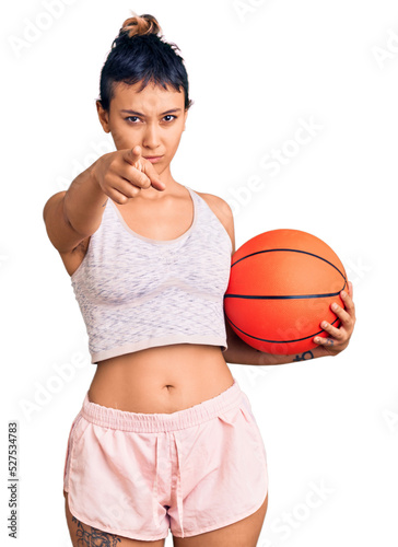 Young woman holding basketball ball pointing with finger to the camera and to you, confident gesture looking serious © Krakenimages.com