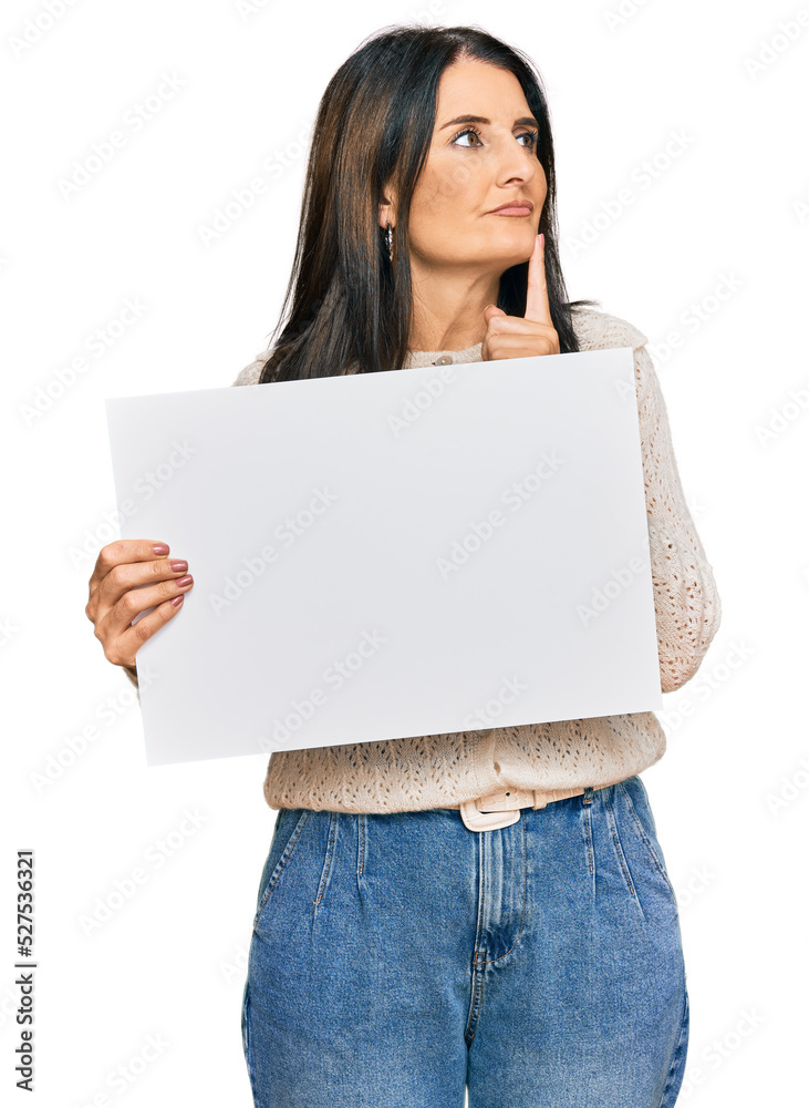 Middle age brunette woman holding blank empty banner serious face thinking about question with hand on chin, thoughtful about confusing idea Middle age brunette woman holding blank empty banner serious face thinking about question with hand on chin, thoughtful about confusing idea