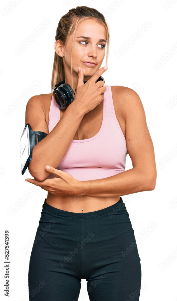 Young blonde woman wearing gym clothes and using headphones with hand on chin thinking about question, pensive expression. smiling with thoughtful face. doubt concept. Young blonde woman wearing gym clothes and using headphones with hand on chin thinking about question, pensive expression. smiling with thoughtful face. doubt concept.
