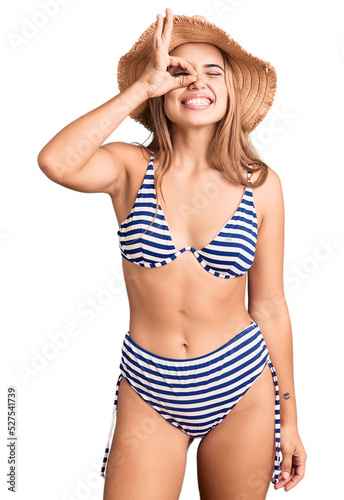 Young beautiful blonde woman wearing bikini and hat doing ok gesture with hand smiling, eye looking through fingers with happy face. photo