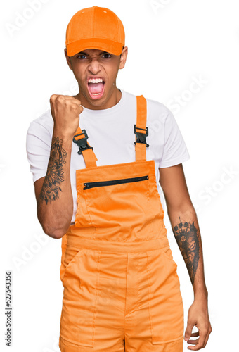 Young handsome african american man wearing handyman uniform angry and mad raising fist frustrated and furious while shouting with anger. rage and aggressive concept.