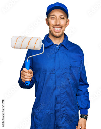 Fototapeta Naklejka Na Ścianę i Meble -  Bald man with beard holding roller painter looking positive and happy standing and smiling with a confident smile showing teeth