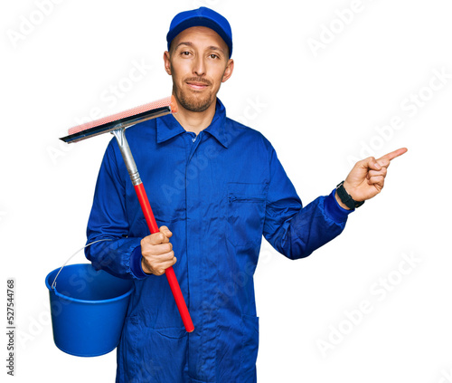 Bald man with beard wearing glass cleaner uniform and squeegee smiling happy pointing with hand and finger to the side