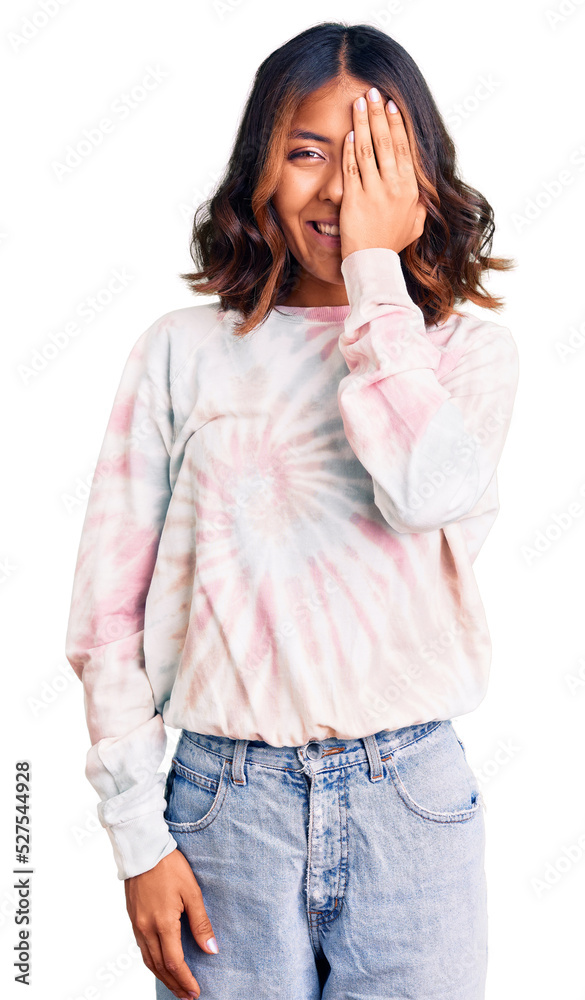 Young beautiful mixed race woman wearing casual tie dye sweatshirt covering one eye with hand, confident smile on face and surprise emotion. Young beautiful mixed race woman wearing casual tie dye sweatshirt covering one eye with hand, confident smile on face and surprise emotion.