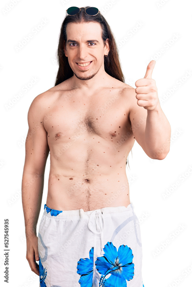 Fototapeta premium Young adult man with long hair wearing swimwear shirtless smiling happy and positive, thumb up doing excellent and approval sign