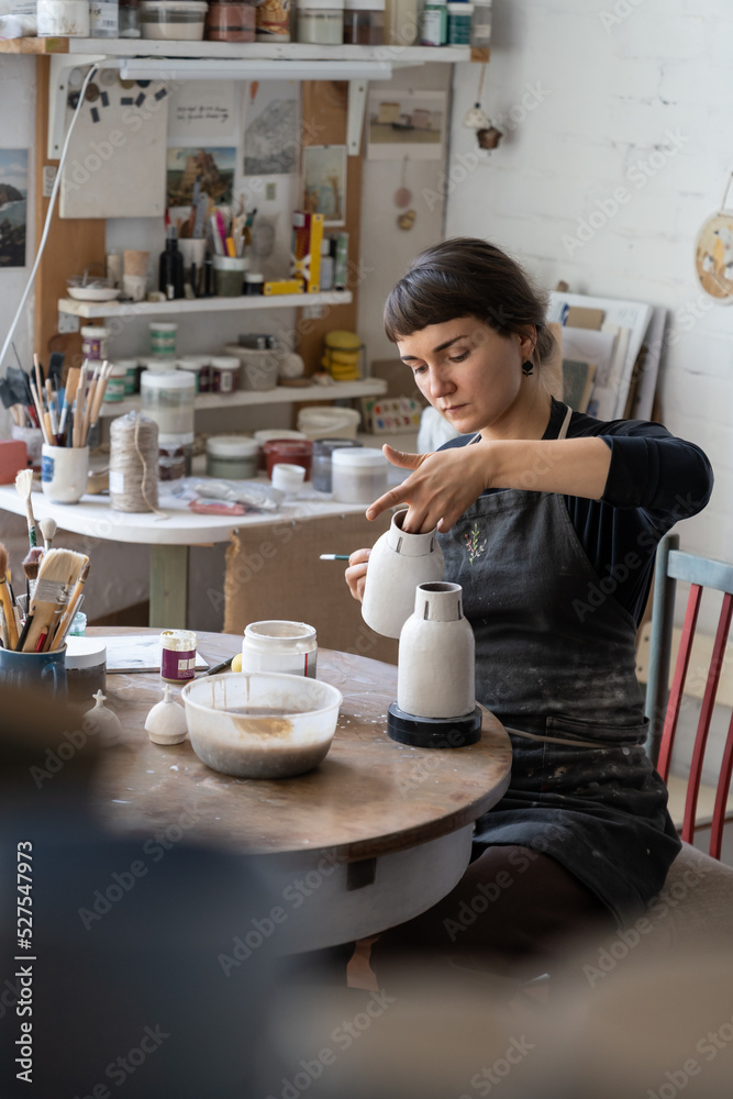 Brunette lady in black apron enjoys new creative hobby learning to work with pottery craft. Focused woman sits at round wooden table painting white handmade crockery with paintbrush in workshop Brunette lady in black apron enjoys new creative hobby learning to work with pottery craft. Focused woman sits at round wooden table painting white handmade crockery with paintbrush in workshop