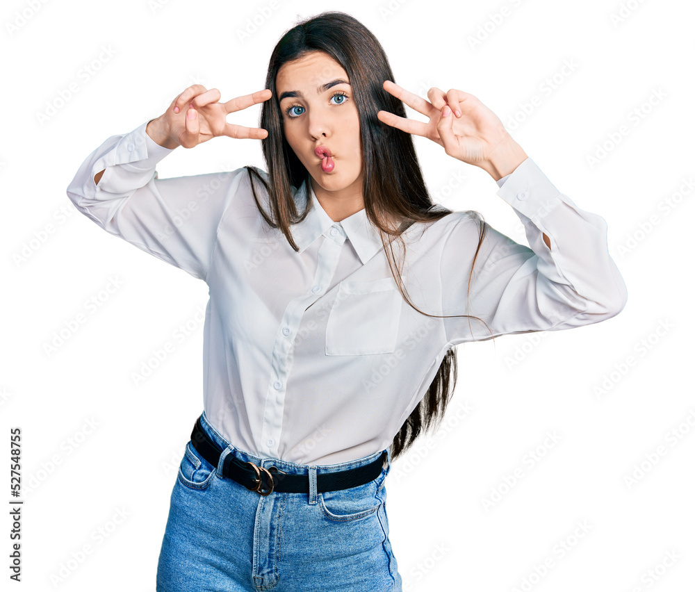 Young brunette teenager doing peace gesture close to eyes making fish face with mouth and squinting eyes, crazy and comical. Young brunette teenager doing peace gesture close to eyes making fish face with mouth and squinting eyes, crazy and comical.