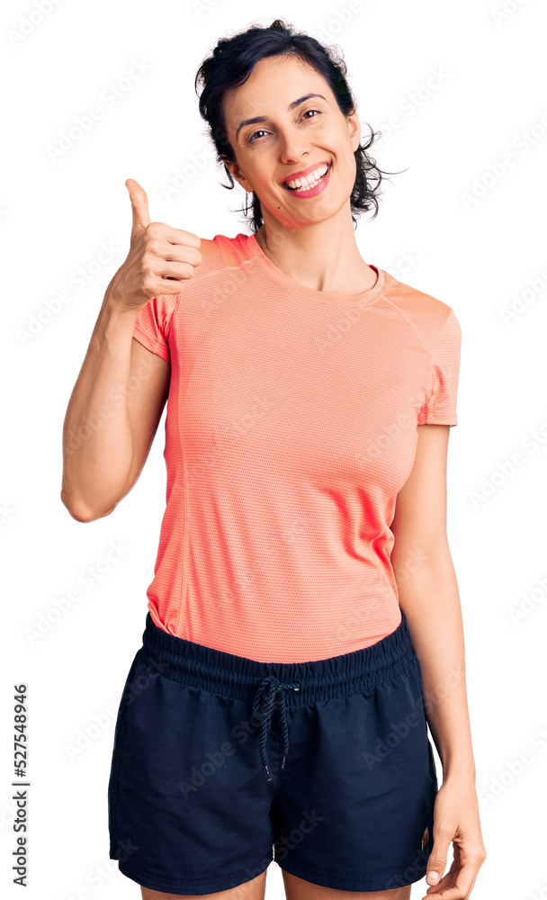 Young beautiful hispanic woman wearing sportswear smiling happy and positive, thumb up doing excellent and approval sign Young beautiful hispanic woman wearing sportswear smiling happy and positive, thumb up doing excellent and approval sign