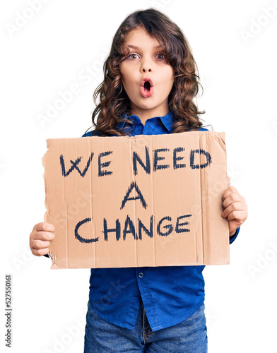 Cute hispanic child with long hair holding we need a change banner scared and amazed with open mouth for surprise, disbelief face