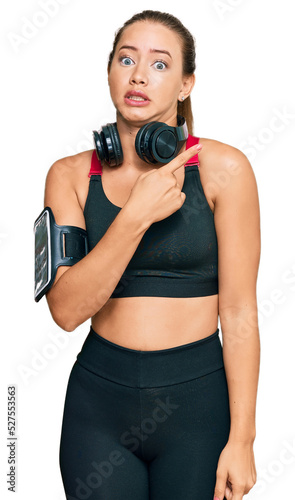 Beautiful blonde woman wearing gym clothes and using headphones pointing aside worried and nervous with forefinger, concerned and surprised expression