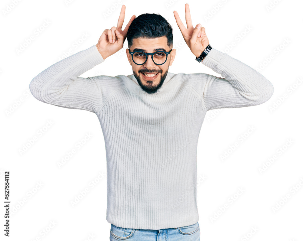 Fototapeta premium Young arab man with beard wearing elegant turtleneck sweater and glasses posing funny and crazy with fingers on head as bunny ears, smiling cheerful