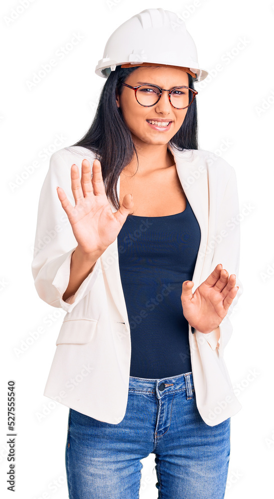 Young beautiful latin girl wearing architect hardhat and glasses disgusted expression, displeased and fearful doing disgust face because aversion reaction. with hands raised Young beautiful latin girl wearing architect hardhat and glasses disgusted expression, displeased and fearful doing disgust face because aversion reaction. with hands raised