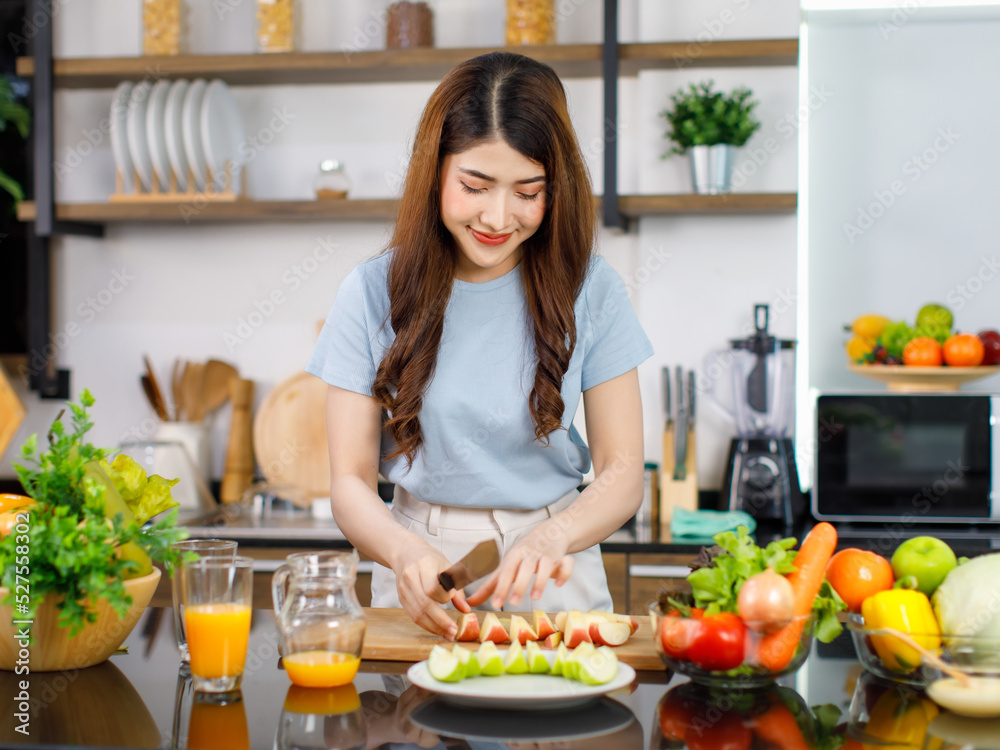 Asian young beautiful housewife standing at kitchen counter full of organic fresh fruits and vegetables bowl using knife preparing cutting red and green apple on chopping board ready to serve on dish Asian young beautiful housewife standing at kitchen counter full of organic fresh fruits and vegetables bowl using knife preparing cutting red and green apple on chopping board ready to serve on dish