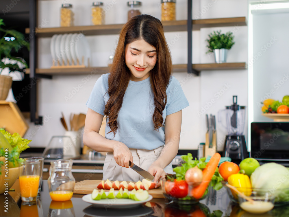 Asian young beautiful housewife standing at kitchen counter full of organic fresh fruits and vegetables bowl using knife preparing cutting red and green apple on chopping board ready to serve on dish