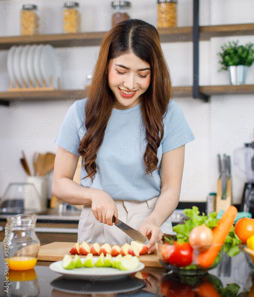 Asian young beautiful housewife standing at kitchen counter full of organic fresh fruits and vegetables bowl using knife preparing cutting red and green apple on chopping board ready to serve on dish