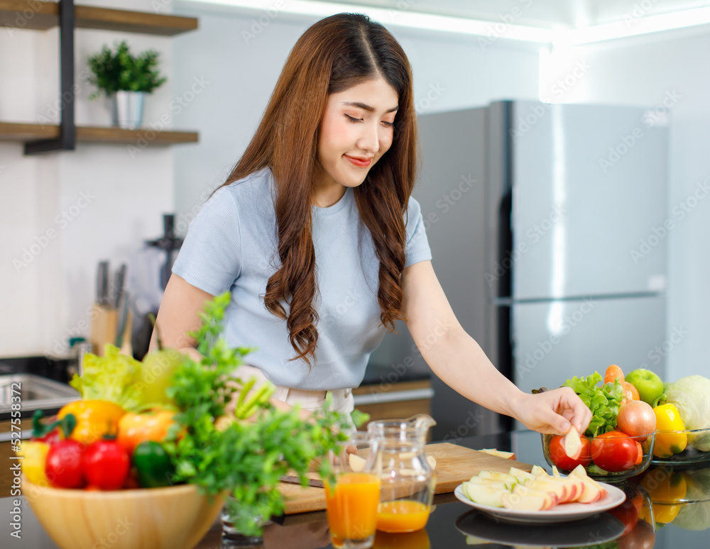 Asian young beautiful housewife standing at kitchen counter full of organic fresh fruits and vegetables bowl using knife preparing cutting red and green apple on chopping board ready to serve on dish Asian young beautiful housewife standing at kitchen counter full of organic fresh fruits and vegetables bowl using knife preparing cutting red and green apple on chopping board ready to serve on dish