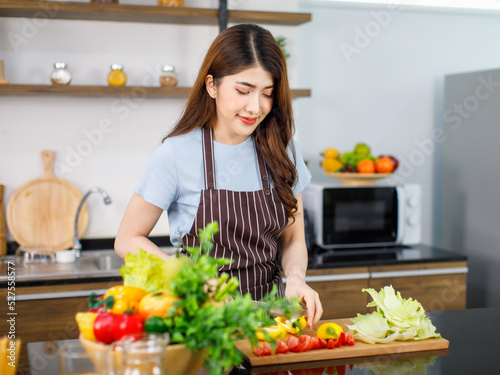 Asian young beautiful housewife in stripe apron standing smiling at kitchen counter full of organic fresh fruits and vegetables in bowls preparing salad peeling lettuce with hands on chopping board