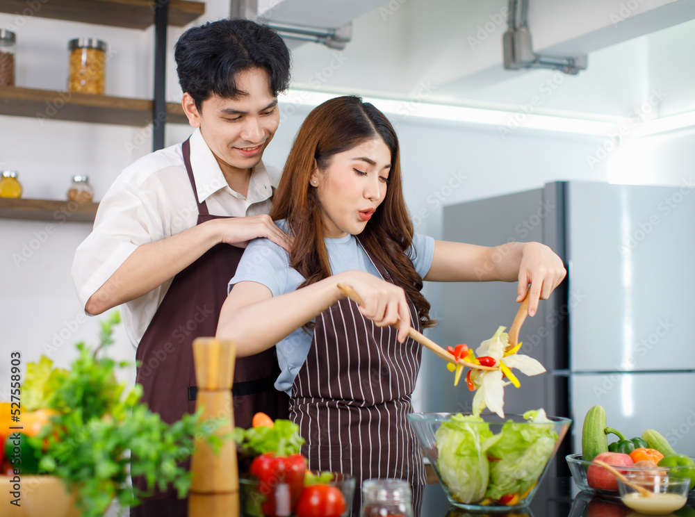 Asian young cheerful beautiful housewife in stripe apron standing smiling at kitchen counter using wood utensils mixing organic fresh salad vegetables in bowl while husband help adding cream dressing Asian young cheerful beautiful housewife in stripe apron standing smiling at kitchen counter using wood utensils mixing organic fresh salad vegetables in bowl while husband help adding cream dressing