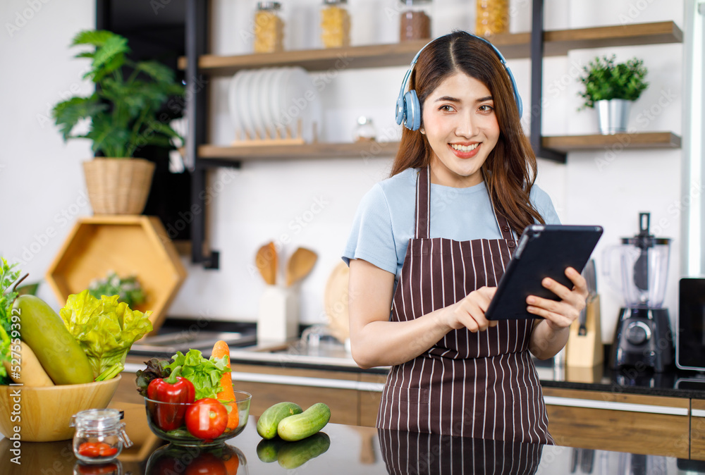 Portrait shot Asian young cheerful housewife in apron wearing wireless headphones listening to online streaming music playlist from touchscreen tablet computer standing smiling behind kitchen counter