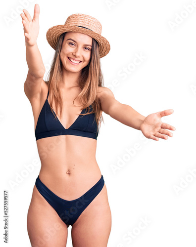 Young beautiful girl wearing bikini and hat looking at the camera smiling with open arms for hug. cheerful expression embracing happiness.