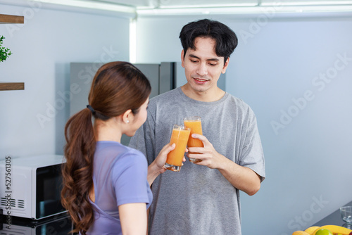 Asian young romantic lover couple handsome husband and beautiful wife standing smiling holding glasses of orange juice toasting cheers together behind kitchen counter full with fresh raw fruits bowls