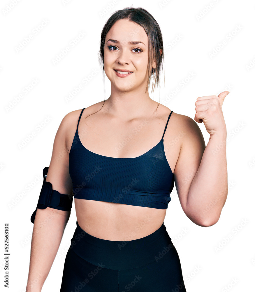 Young hispanic woman wearing sportswear smiling with happy face looking and pointing to the side with thumb up. Young hispanic woman wearing sportswear smiling with happy face looking and pointing to the side with thumb up.