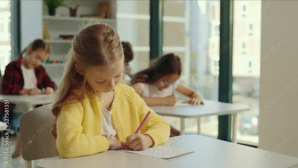 Beautiful long-haired blonde pupil sits at a first desk in the classroom and fills out tests thinks about answers, and the pupils doing test on the background. Back to school. Beautiful long-haired blonde pupil sits at a first desk in the classroom and fills out tests thinks about answers, and the pupils doing test on the background. Back to school.