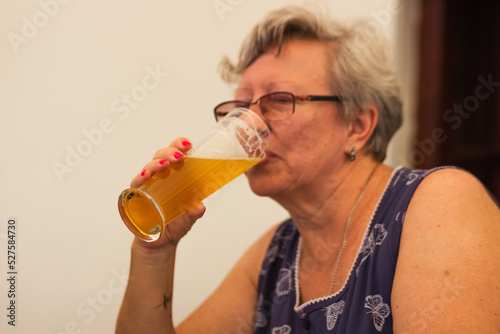 Smiling caucasian senior woman with gray hair holding and drinking glass of light beer in craft czech brewery.