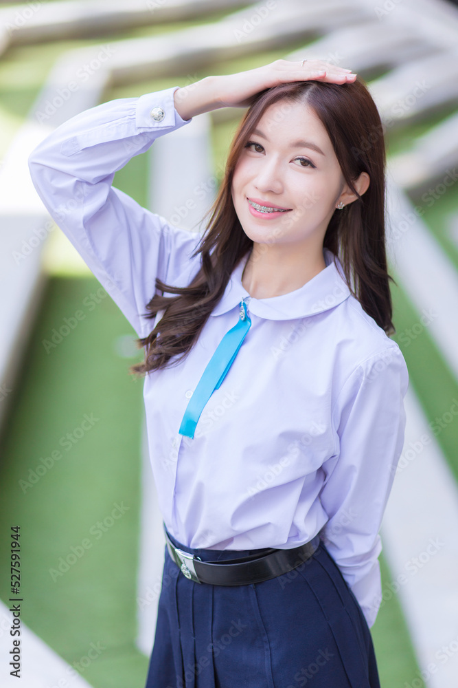 Beautiful high school Asian student girl in the school uniform stands and smiles happily with braces on her teeth while she put her hand on head confidently with the building as a background.