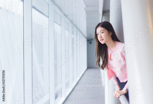 Beautiful teenage Asian girl wears a white with pink shirt and smiles cheerfully and confident while she stands near the glassed window on a nice day.