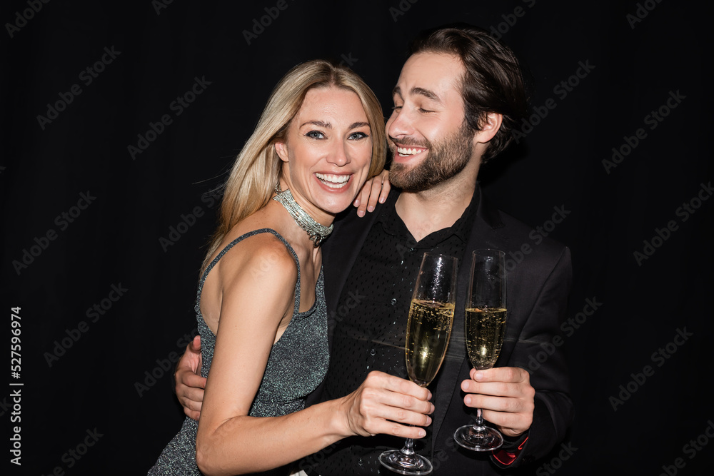 Bearded man hugging cheerful girlfriend with glass of champagne isolated on black.