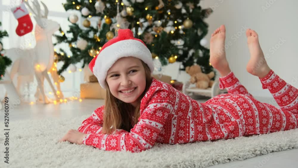 Happy child girl lying on floor near to Christmas tree wearing red Santa hat and smiling. Beautiful female kid enjoying New Year and Xmas time