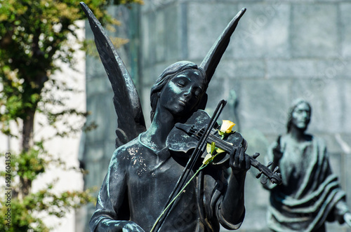 La statua in bronzo di un angelo che suona il violino su cui hanno appoggiato una rosa gialla su una tomba dell cimitero maggiore di Milano photo