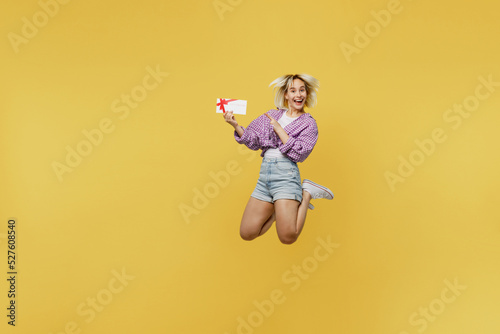 Full body young excited blonde woman 20s she wear pink tied shirt white t-shirt jump high hold point index finger on gift certificate coupon voucher card for store isolated on plain yellow background.
