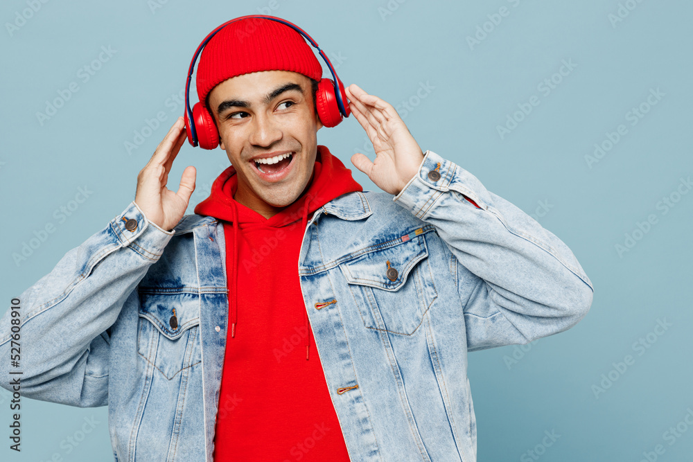Young happy middle eastern man wear denim jacket red hat headphones listen to music look aside on workspace area mock up isolated on plain pastel light blue cyan background. People lifestyle concept. Young happy middle eastern man wear denim jacket red hat headphones listen to music look aside on workspace area mock up isolated on plain pastel light blue cyan background. People lifestyle concept.