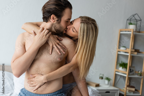 passionate blonde woman embracing and kissing shirtless man in bedroom.