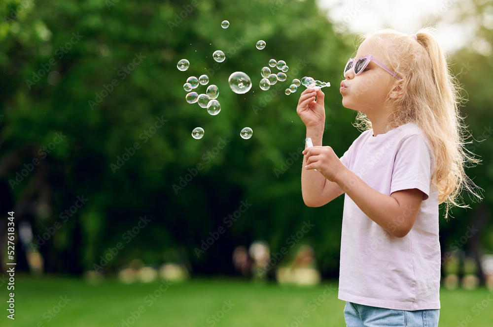 Cheerful caucasian blond preschool girl wearing sunglasses blowing soap bubbles in summer park outdoors and have fun. Happy childhood concept